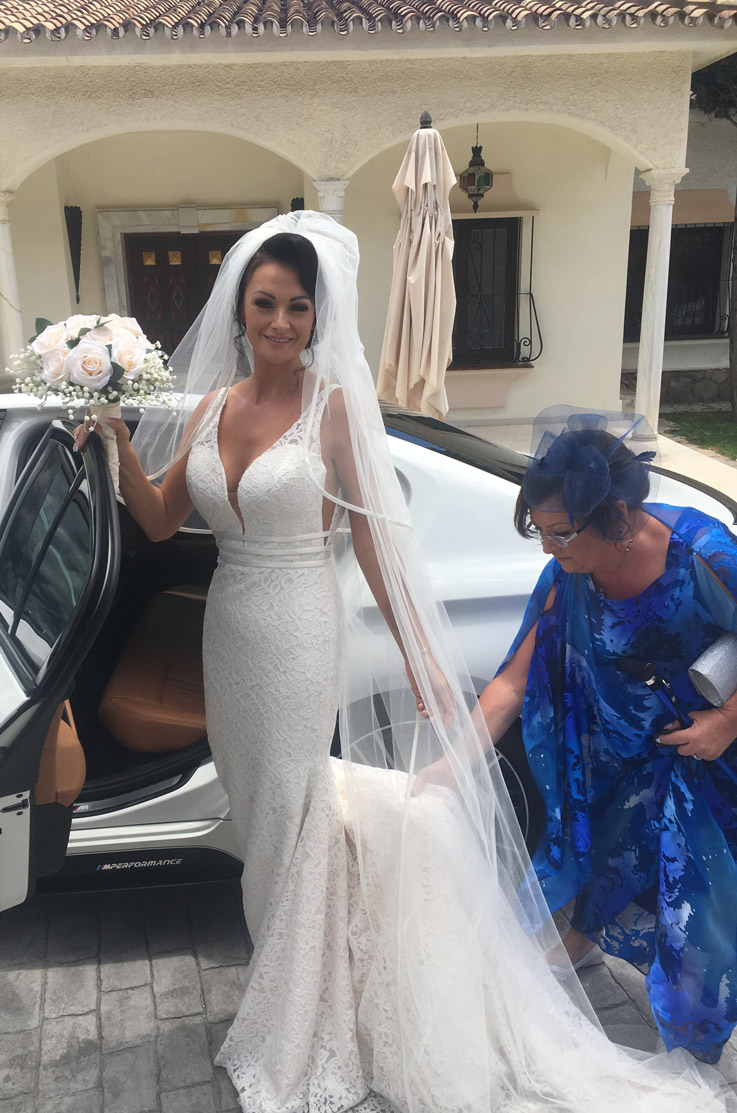 Bride exits a white car in a lace plunging neck gown with a flowing veil, and is holding a bouquet in front of the venue