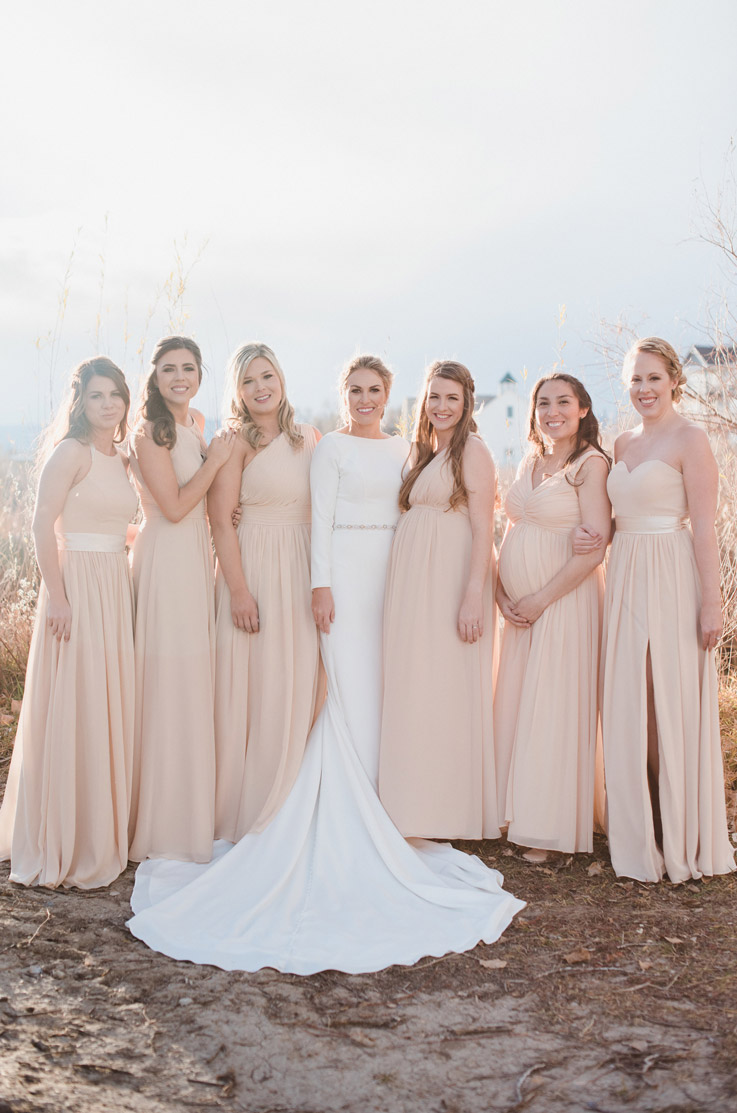 Bride in a high neck fit and flare gown with sleeves and a crepe belt, stands next to bridesmaids in light pink outdoors.