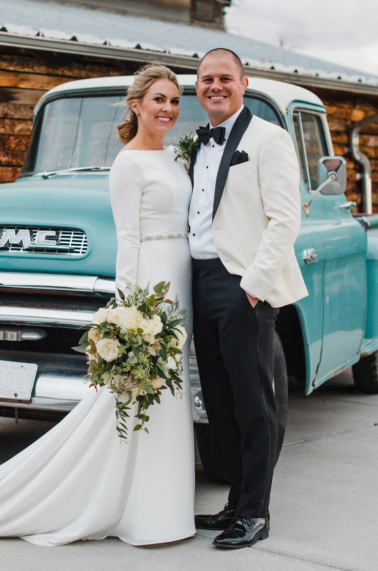 Bride stands next to her groom in a crepe long-sleeve gown with belt detail, holding a bouquet in front of a blue vintage car.