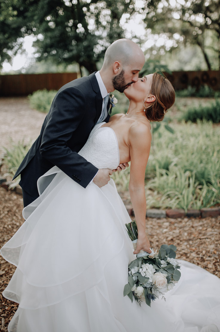 Groom kisses his bride in a strapless ballgown with a tiered skirt and lace bodice, and is holding a bouquet outdoors.