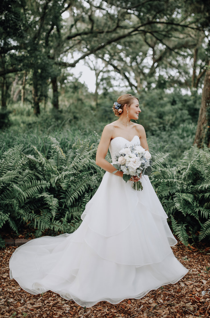 Bride in an outdoor setting surrounded by leaves, wearing a strapless organza gown with a tiered skirt and chapel train.