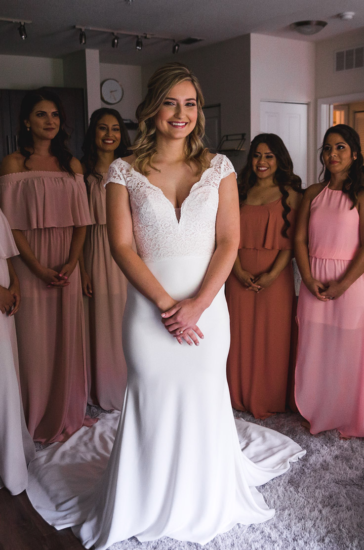  Bride poses in front of bridesmaids in pink dresses, in a lace fit and flare gown with a plunging neck and cap sleeves, indoors.