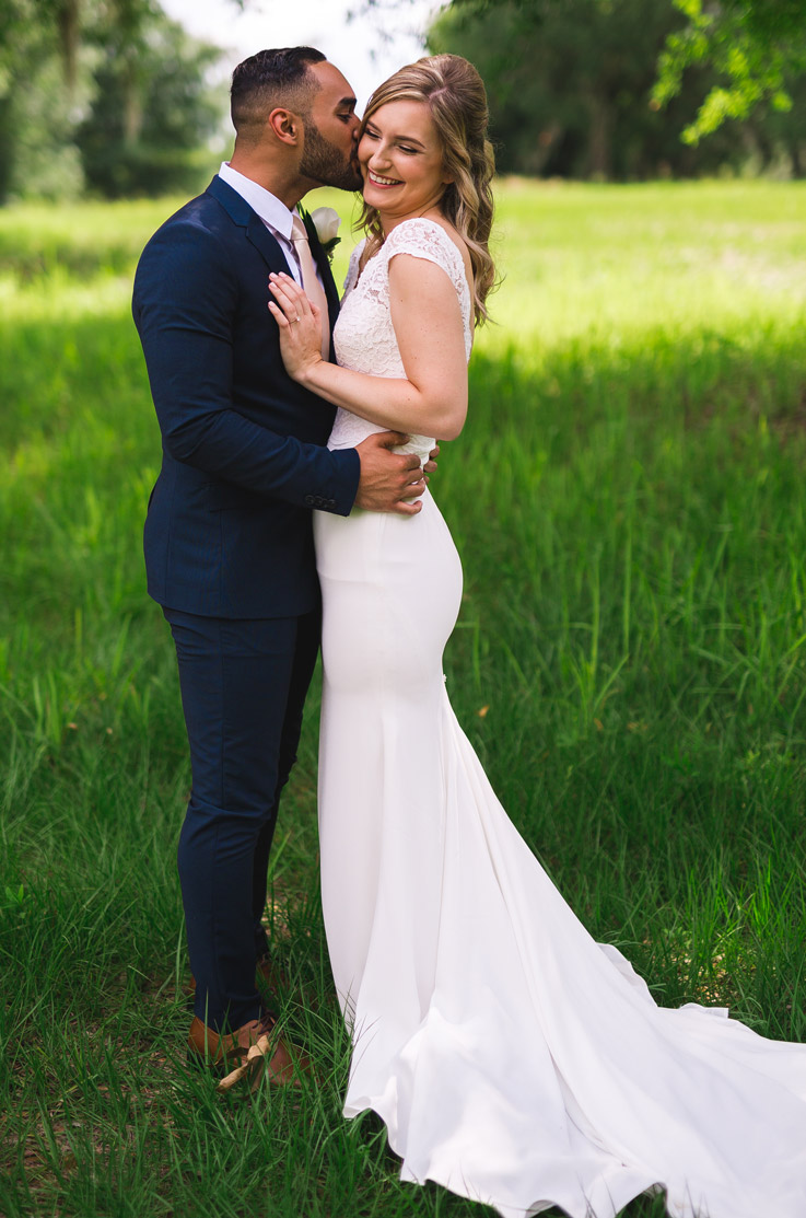 Groom embraces bride in a lace fit and flare gown with cap sleeves and a plunging neckline as they stand in a grassy field.