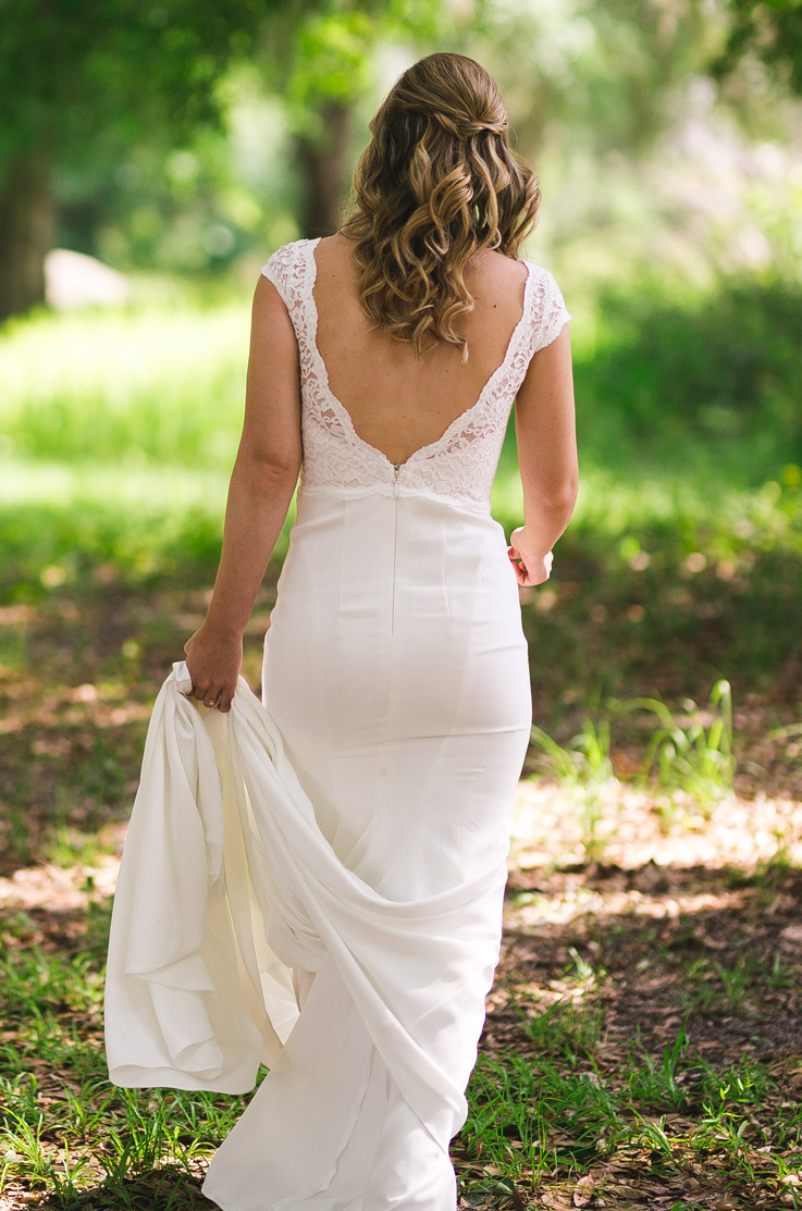 Bride in a deep-V back lace wedding gown with scalloping around the hem, holds the end of the train in the grass.