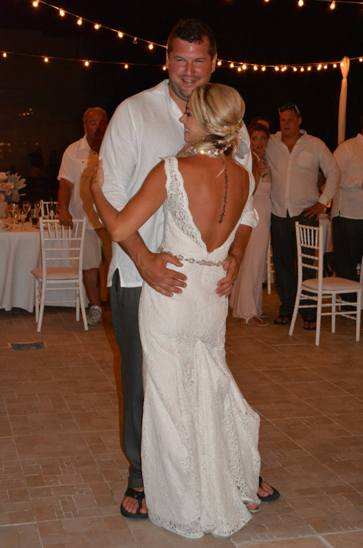 Bride in a lace open-back wedding dress with a scalloped neckline and beaded belt, dances with groom at reception under lights.