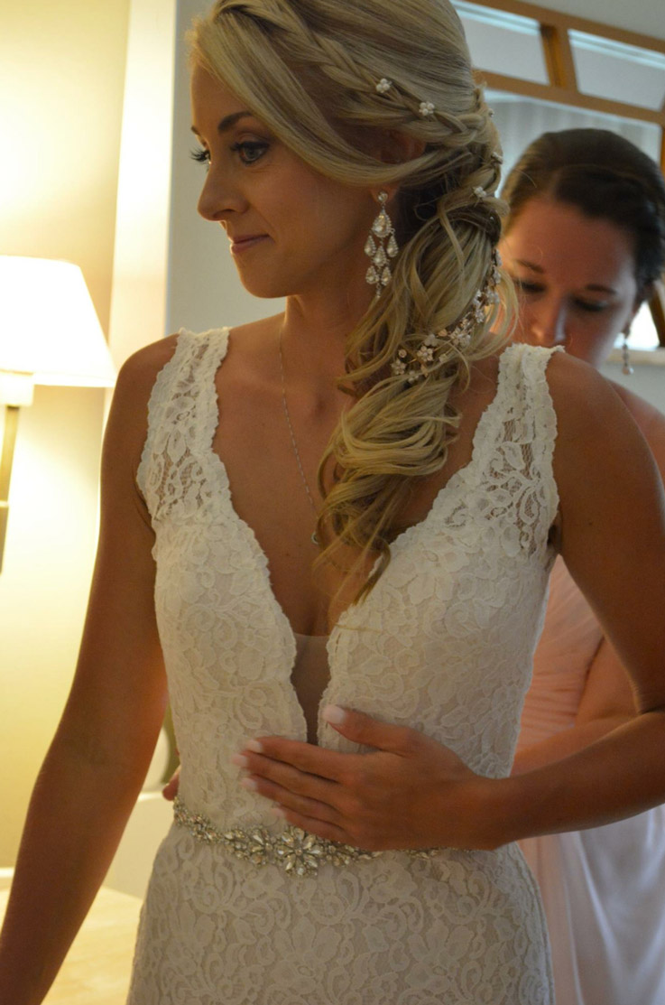 Close-up of bride getting ready in a bridal suite, wearing a plunging neck lace gown with a beaded band and is fastened into it.