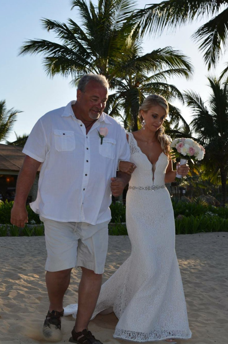 Bride in a lace fit and flare gown with a beaded band and plunging neck, walks with her father on a sandy beach with palm trees.