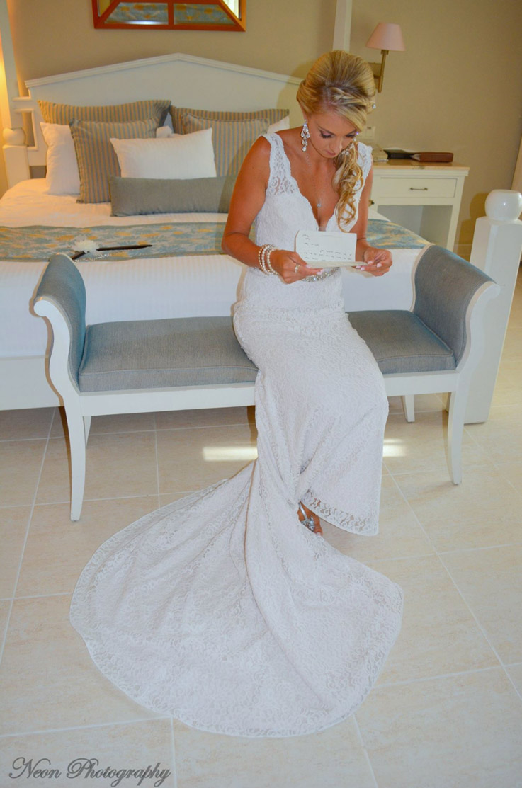 Bride sits on a lounge chair in hotel suite and reads a card, in a plunging neck lace gown with scalloping and a chapel train.