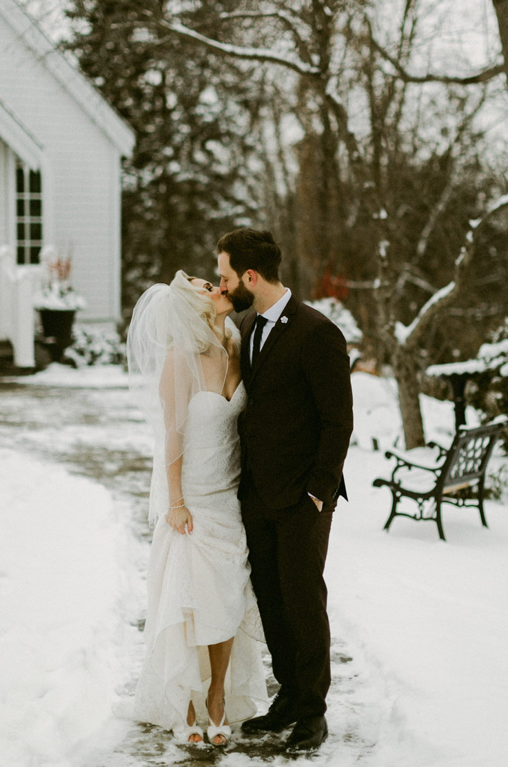 Bride in a lace gown with thin straps and a veil, lifts her train and kisses groom in the snow outside the venue.