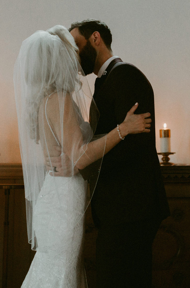 Bride in a fit and flare lace wedding dress with an open back and veil, kisses her groom at intimate candlelit ceremony.