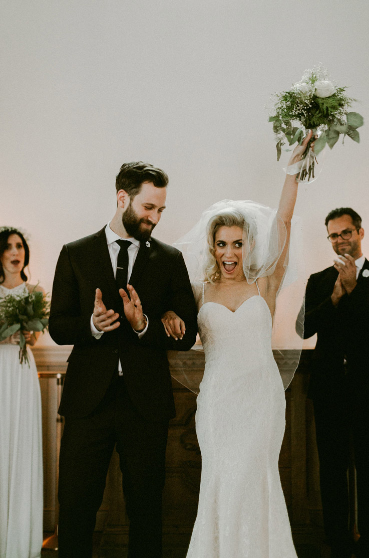 Bride in a spaghetti strap lace gown with a sweetheart neck, lifts her bouquet in celebration, beside the groom in a suit.
