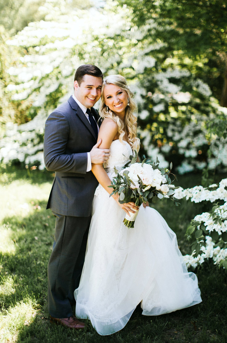 Bride in a strapless lace bodice gown with a tulle overskirt poses with her groom outdoors in a sunlit area with a bouquet.