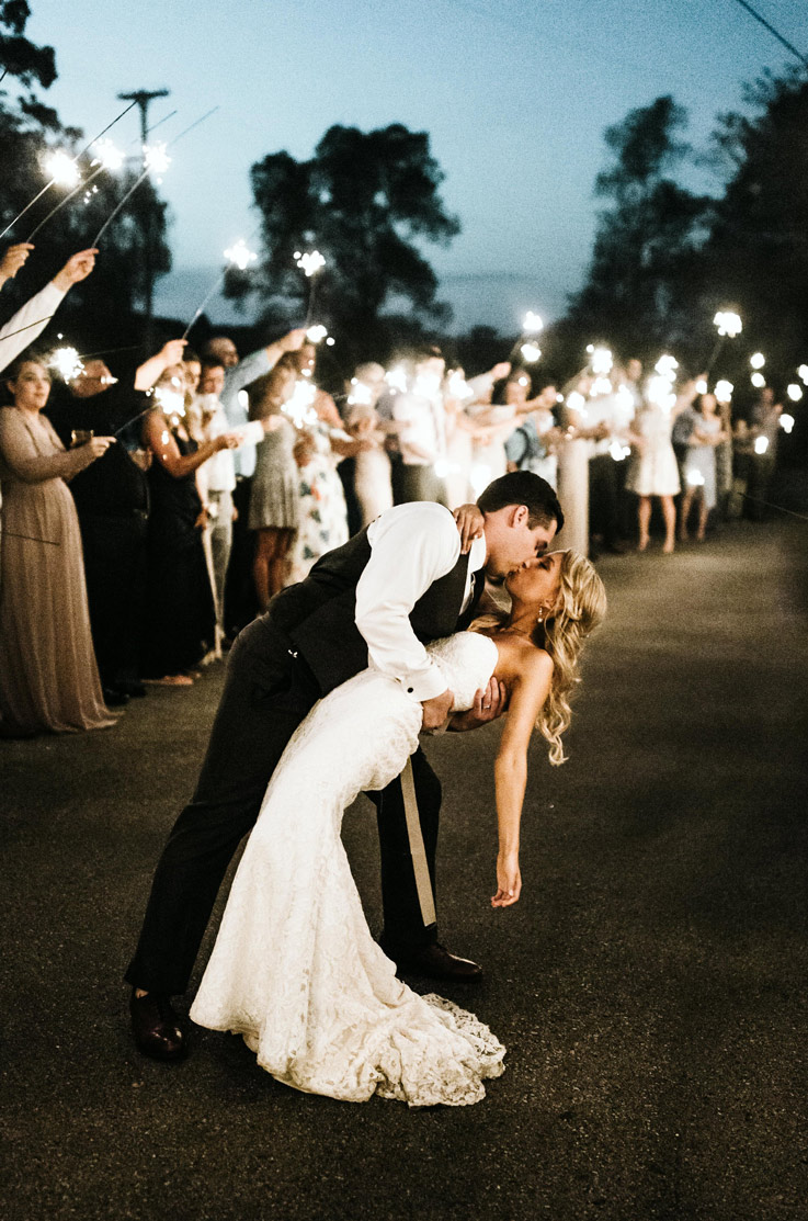 Groom dips the bride in a strapless lace gown with a tulle overskirt and sweetheart neck, in sparkler send-off at night.