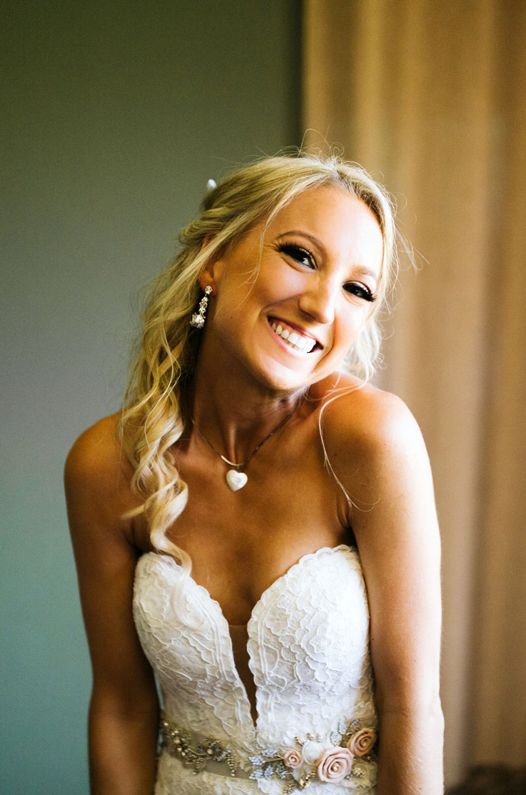 Close-up of a smiling bride in a lace sweetheart neck gown with beaded embellishment at the waist, indoors by a curtain.