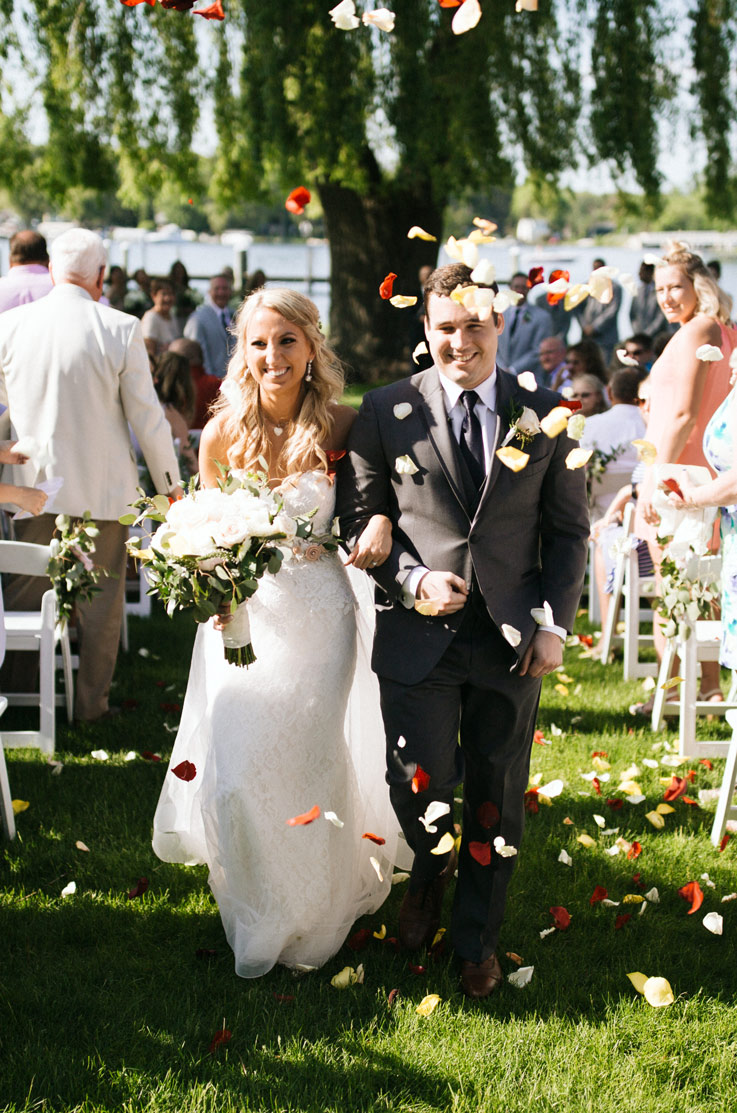 Bride in a lace strapless wedding gown, holding a bouquet and links arms with groom as flower petals fall and they pass guests.
