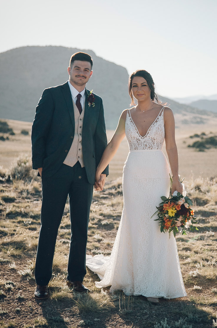 Bride in a V-neck lace wedding dress with a fit and flare skirt, stands in a sunlit desert landscape beside the groom.