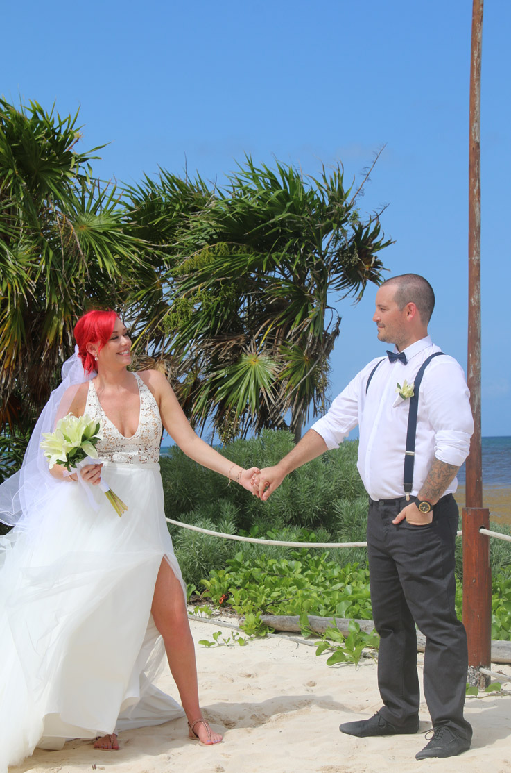 Groom holds hands with bride in a lace bodice gown with a V-neck, and a flowing tulle skirt, standing on a white sand beach.