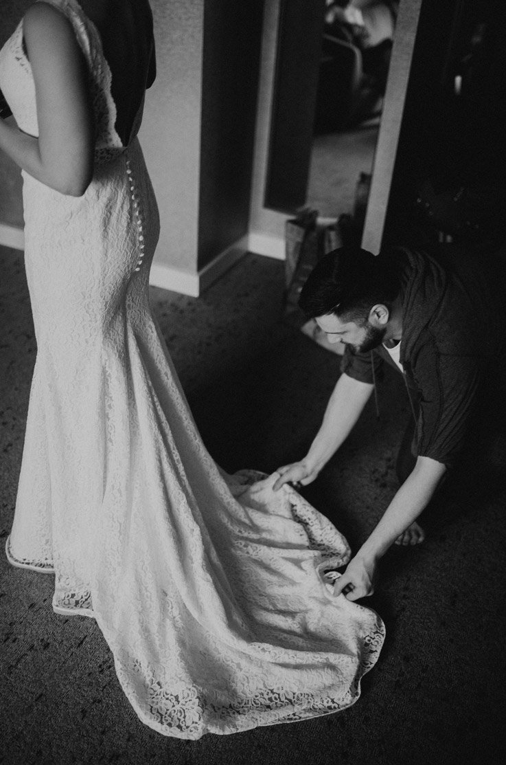 Bride in a low open back fit and flare gown in lace with buttons, stands in a room in black and white, while train gets adjusted.