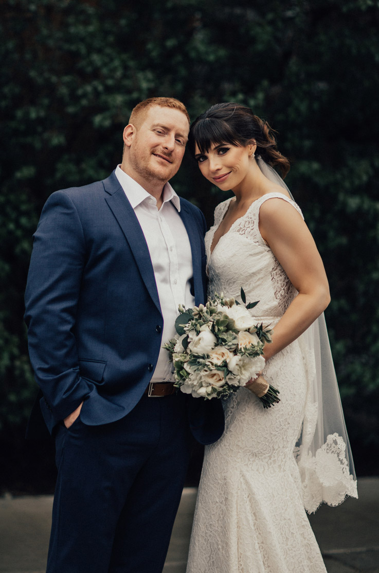 Bride poses next to groom outdoors, in a fit and flare lace wedding dress with a plunging neck and veil, holding a bouquet.