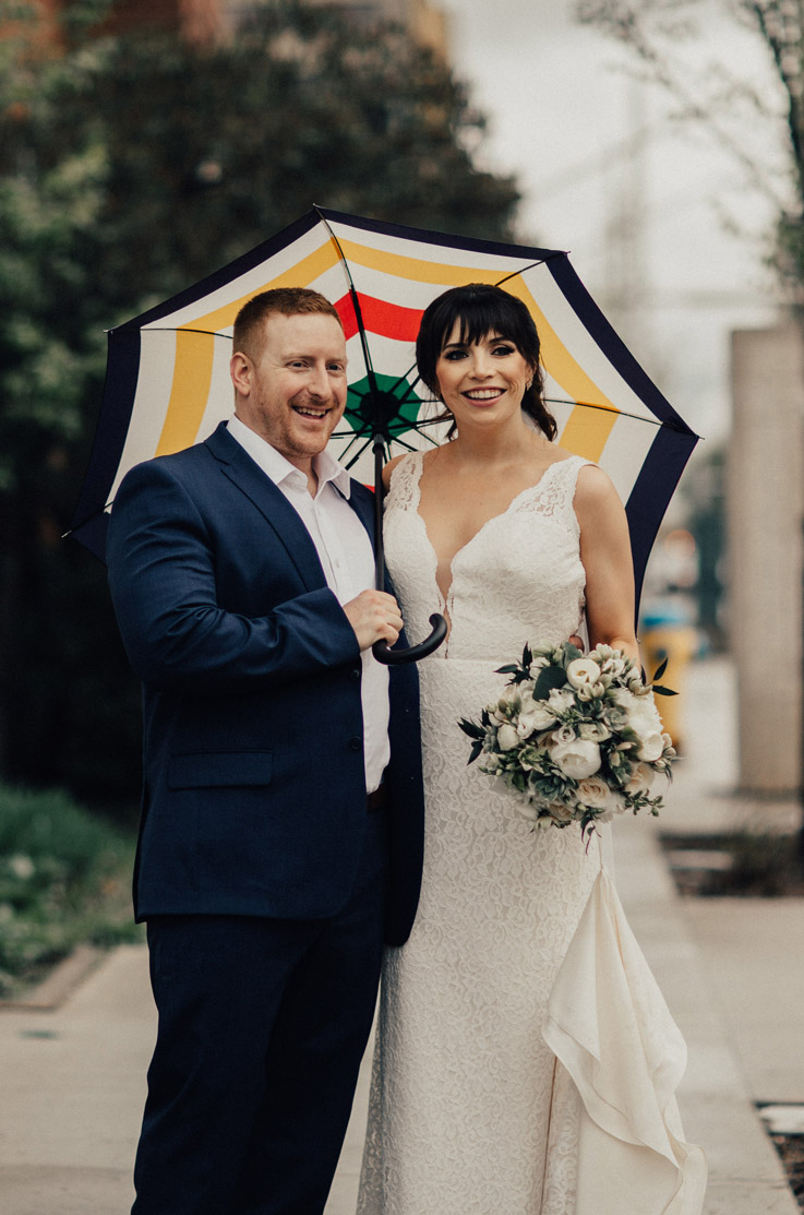 Bride in a lace plunging neck gown with a chapel train, stands beside groom smiling under an umbrella and is holding a bouquet.