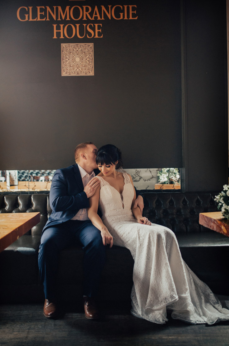 Groom embraces bride in a plunging neck lace wedding gown and they are seated on a leather couch.