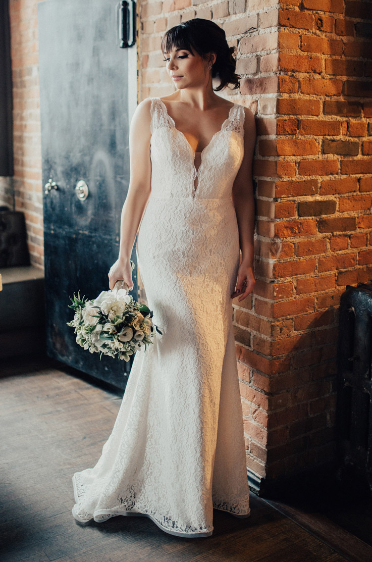 Bride leans against exposed brick wall, in a fit and flare lace gown with a plunging neck, holding a bouquet to the side.