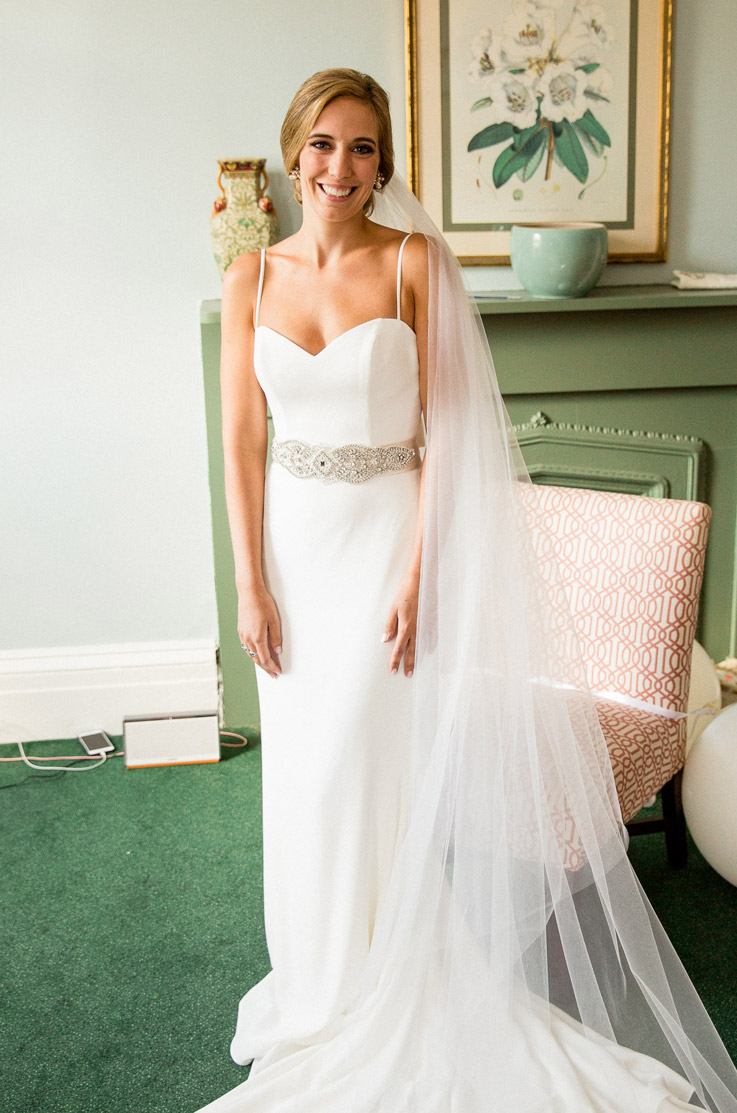 Smiling bride poses in a fit and flare crepe gown with a beaded band, veil and spaghetti straps by a green fireplace.