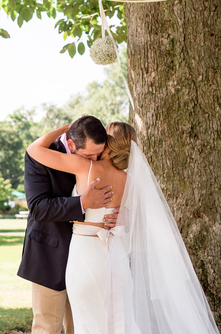 Groom embraces bride under a tree in a spaghetti strap two-piece gown with a veil, crepe skirt and a crop lace top.