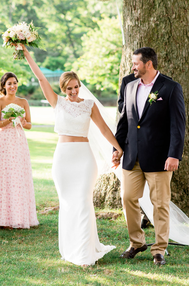 Bride in a two-piece wedding dress with a crop lace top and a high waisted crepe skirt, celebrates outdoors with her groom.
