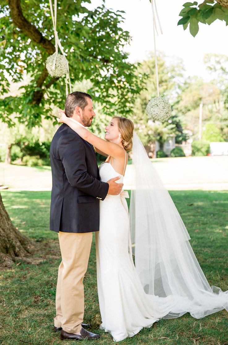 Bride in a wedding dress with lace, a low back, veil and sash, embraces her groom under trees in a sunny garden.