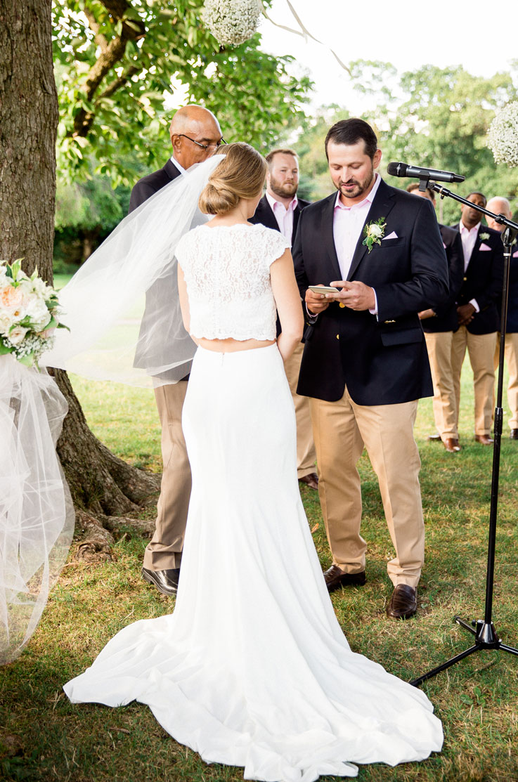 Bride in a two-piece lace gown with a crepe fit and flare skirt and a veil, exchanges vows with the groom in outdoor ceremony.
