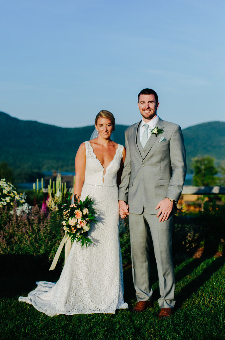 Bride in lace V-neck fit and flare wedding dress, holds a bouquet outdoors and stands with groom on a hilltop.