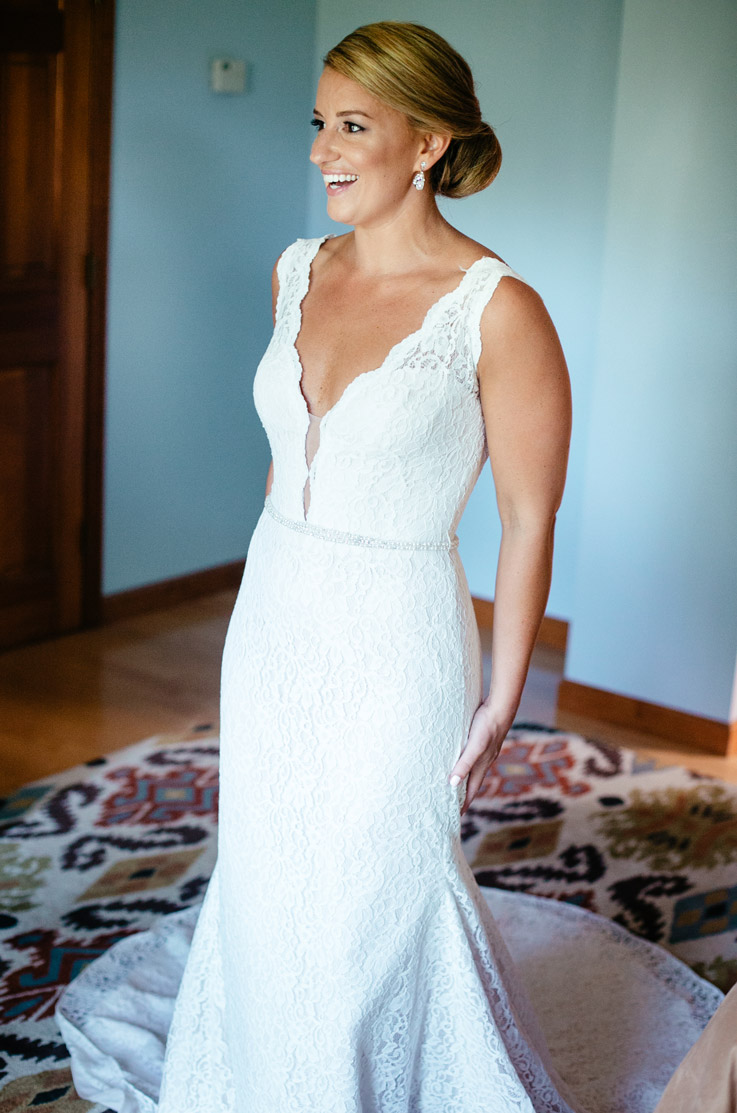 Smiling bride in a lace fit and flare gown with a plunging neck, a thin beaded belt and chapel train, standing indoors.