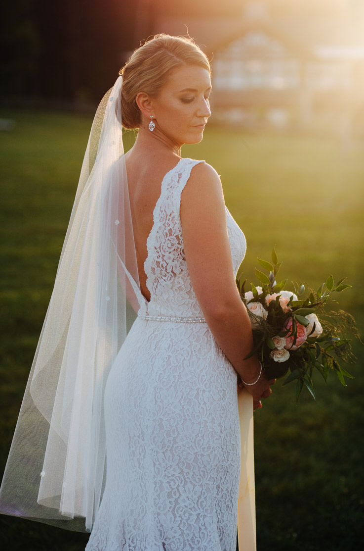 Bride holding a bouquet at sunset, in a deep-V back lace gown with beaded belt detail, a chapel train and flowing veil.