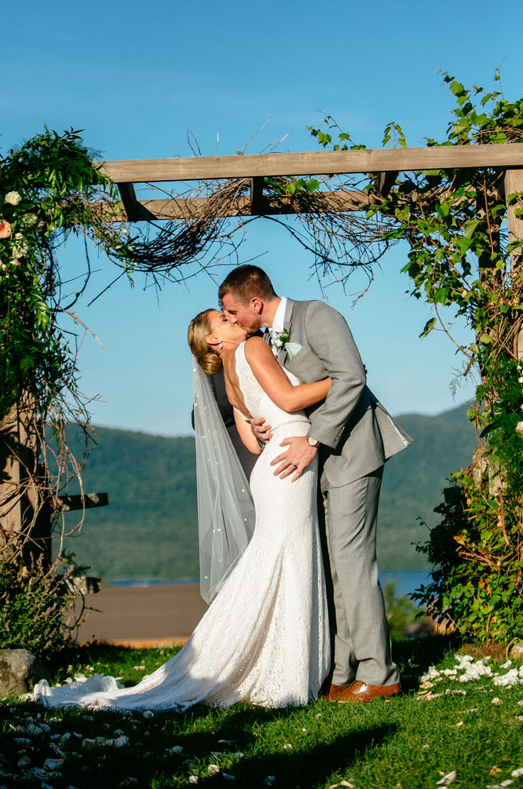 Bride kisses groom in a sleeveless lace wedding gown with a beaded belt and veil, under a rustic wooden arbor.
