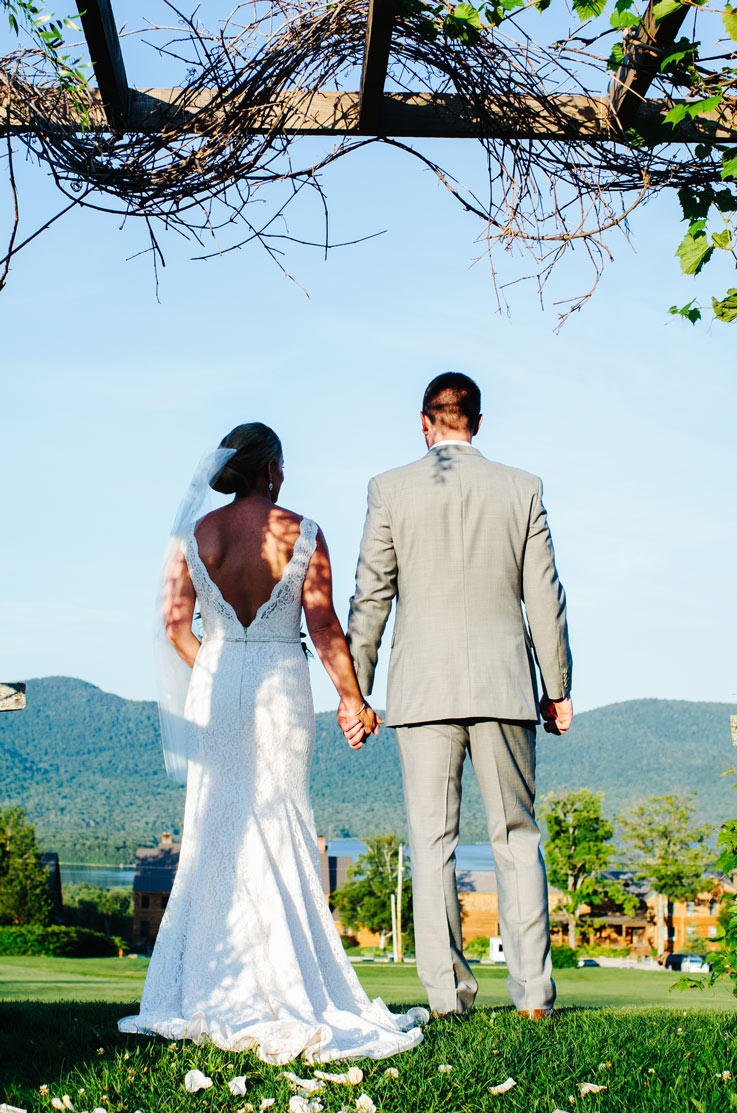 Bride stands on grass, holding hands with groom in a lace fit and flare gown with a low back and veil, looking out at mountains.