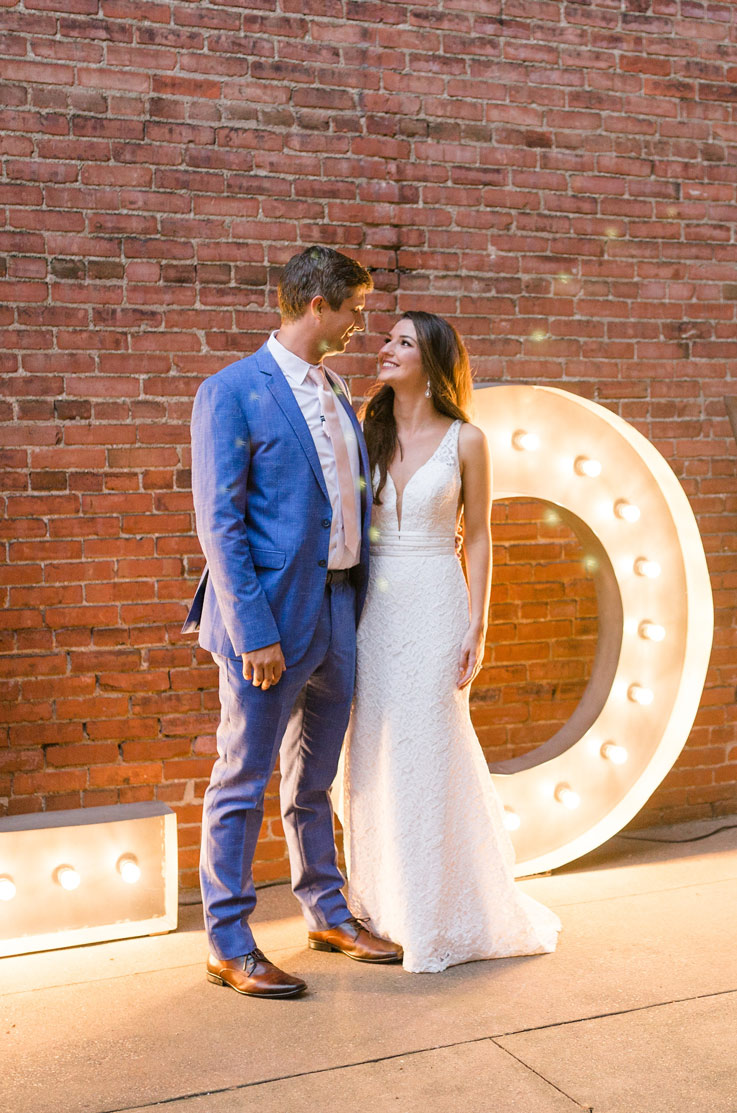 Bride in a fit and flare wedding gown with a plunging neckline, stands with groom by a brick wall and glowing marquee lights.