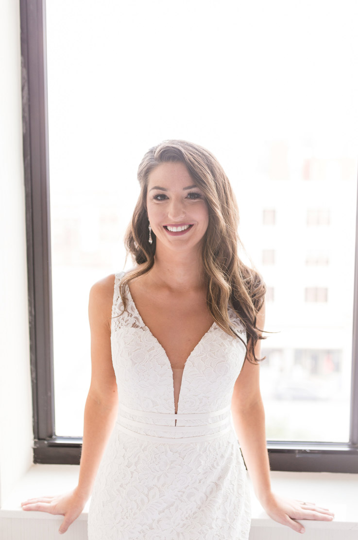 Close-up of a smiling bride in a lace plunging neck gown, with thin waist bands, standing by a bright window.