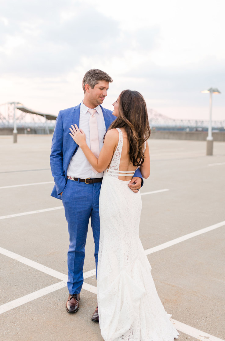 Groom embraces bride in a backless fit and flare gown in lace with thin waistbands, on a rooftop parking lot.