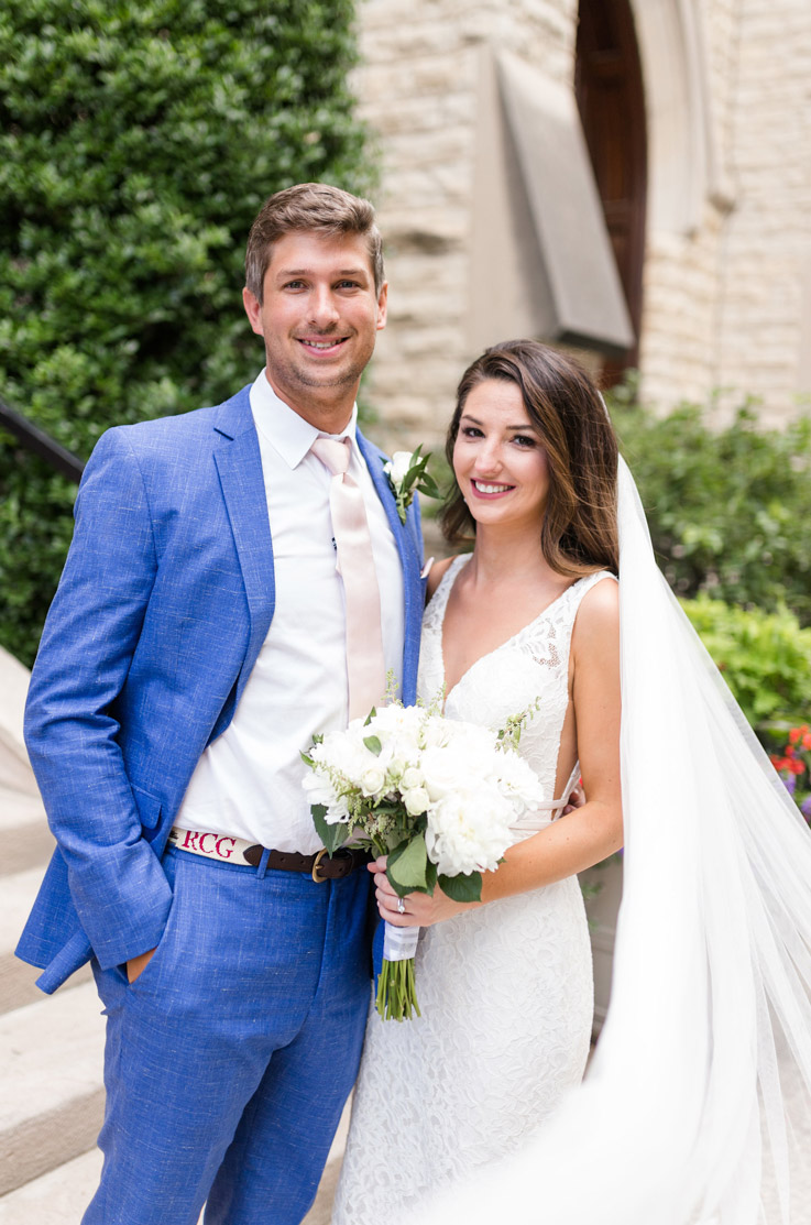 Bride in a plunging neck lace gown with a flowing veil, smiles next to groom in blue suit, outdoors holding a bouquet.