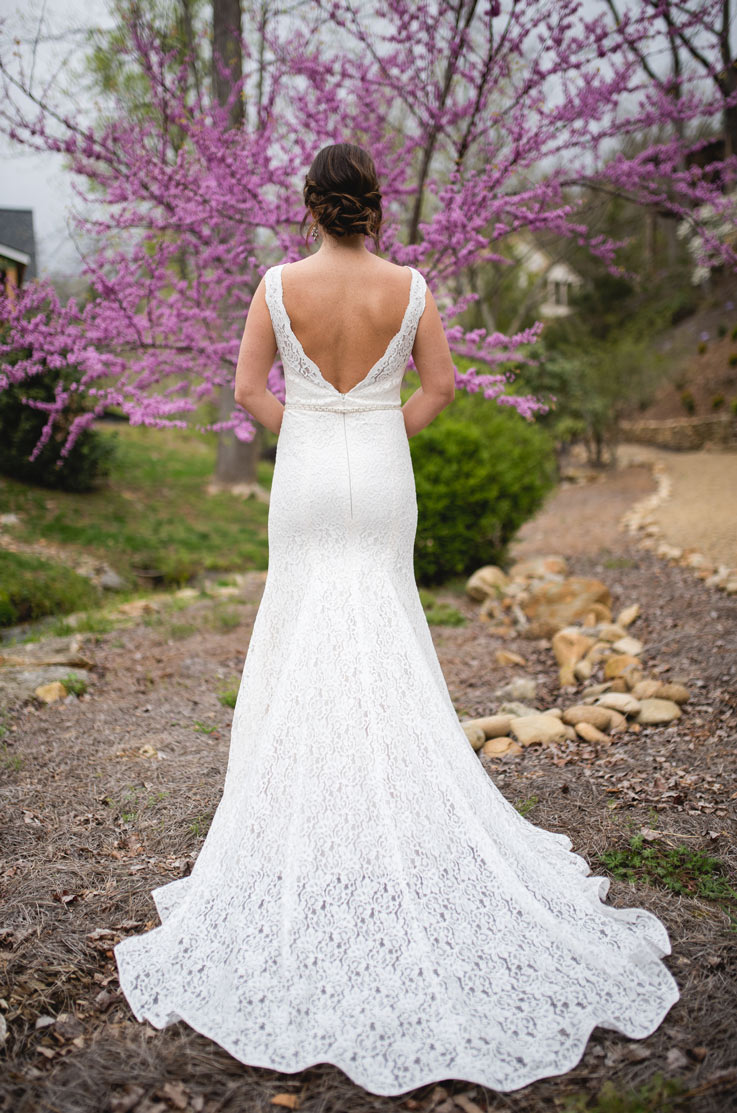 Back of bride in a lace gown with a plunging neckline and chapel train, standing by trees near a path lined with stones.