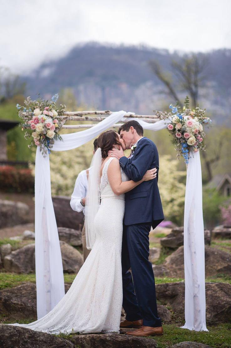 Bride in a lace fit and flare wedding dress with a low back and chapel train, she kisses groom at outdoor mountain ceremony.