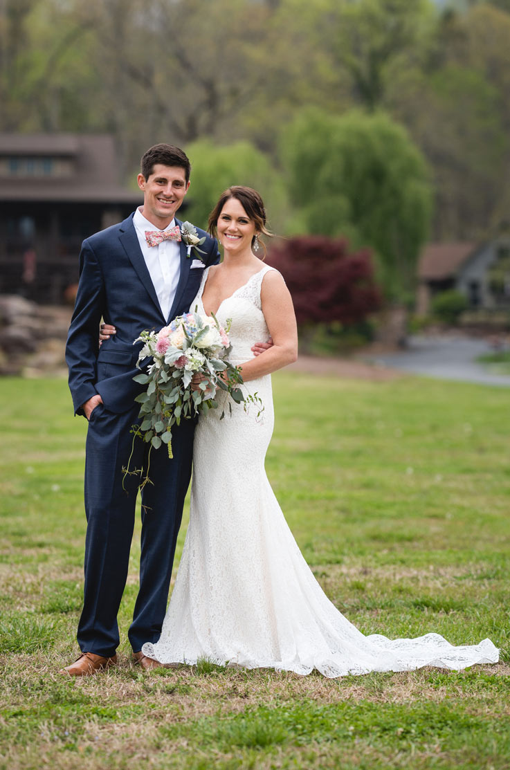 Bride in a plunging neck lace gown with a fit and flare skirt, poses with groom outdoors, and is holding a bouquet.