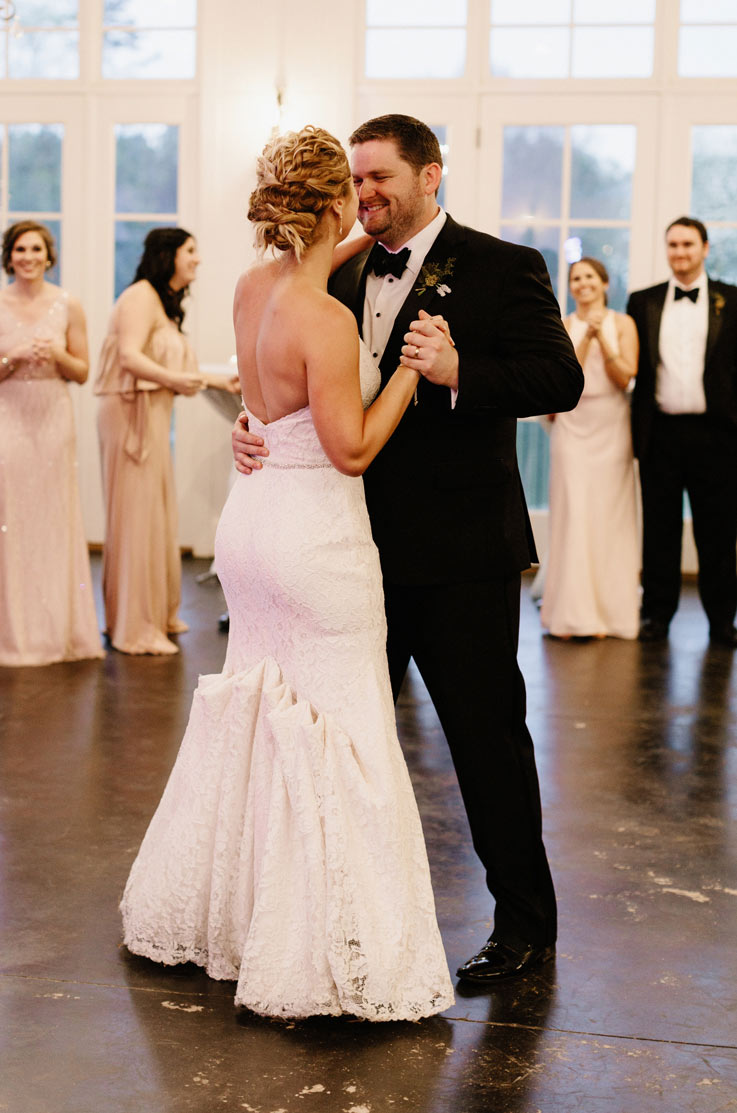 Bride in a strapless lace gown with a fit and flare skirt, dances with groom in a tuxedo at reception, near guests.