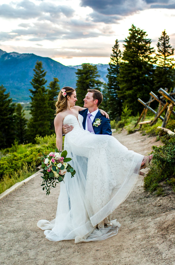 Groom carries his bride in a strapless lace gown with a tulle overskirt, on a mountain overlook spot with pine trees.