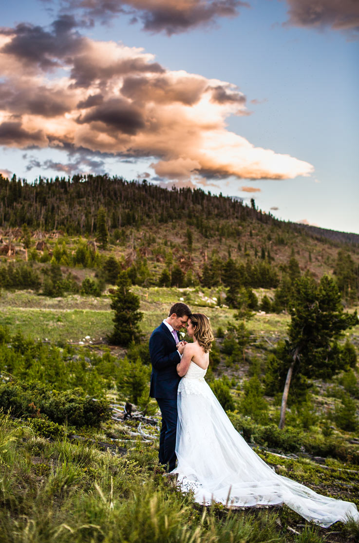 Groom embraces his bride wearing a strapless lace gown with tulle and a low back, standing on a mountain hillside.