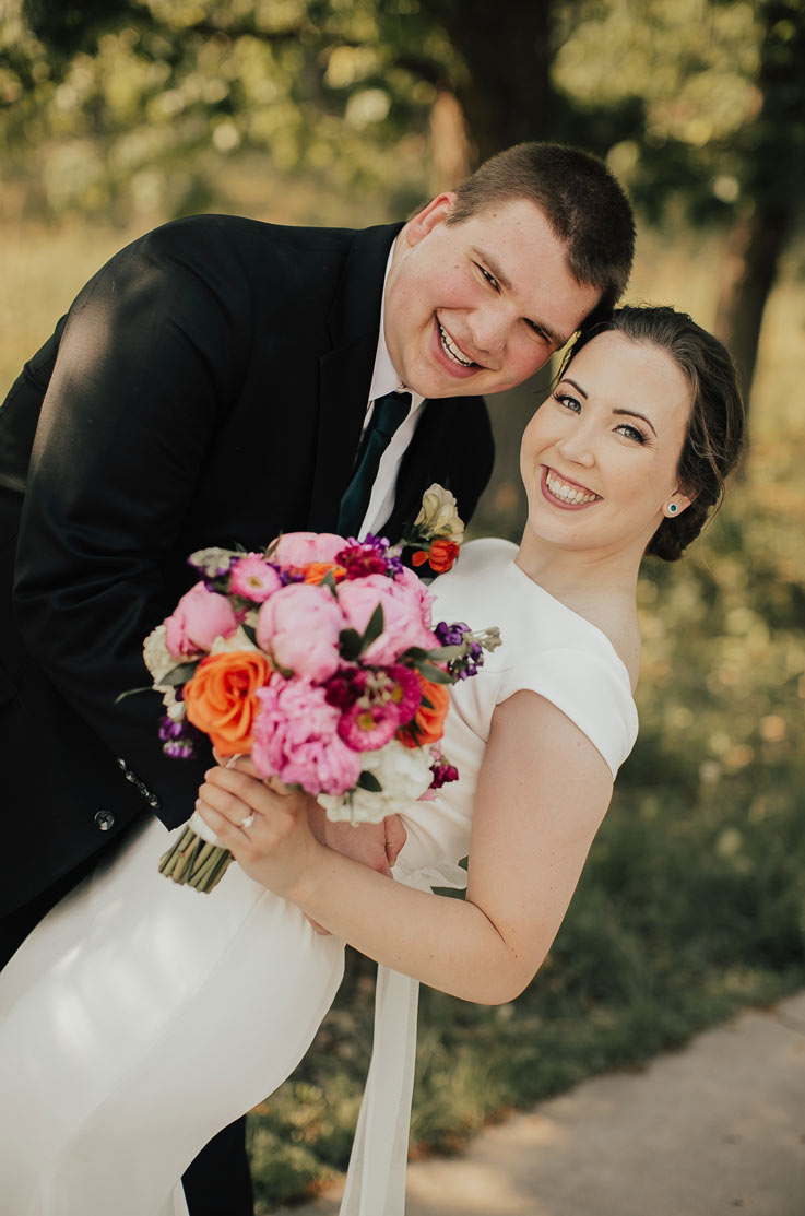 Groom embraces bride in a boat neck gown with cap sleeves, and they are holding a colorful bouquet outdoors near trees. 
