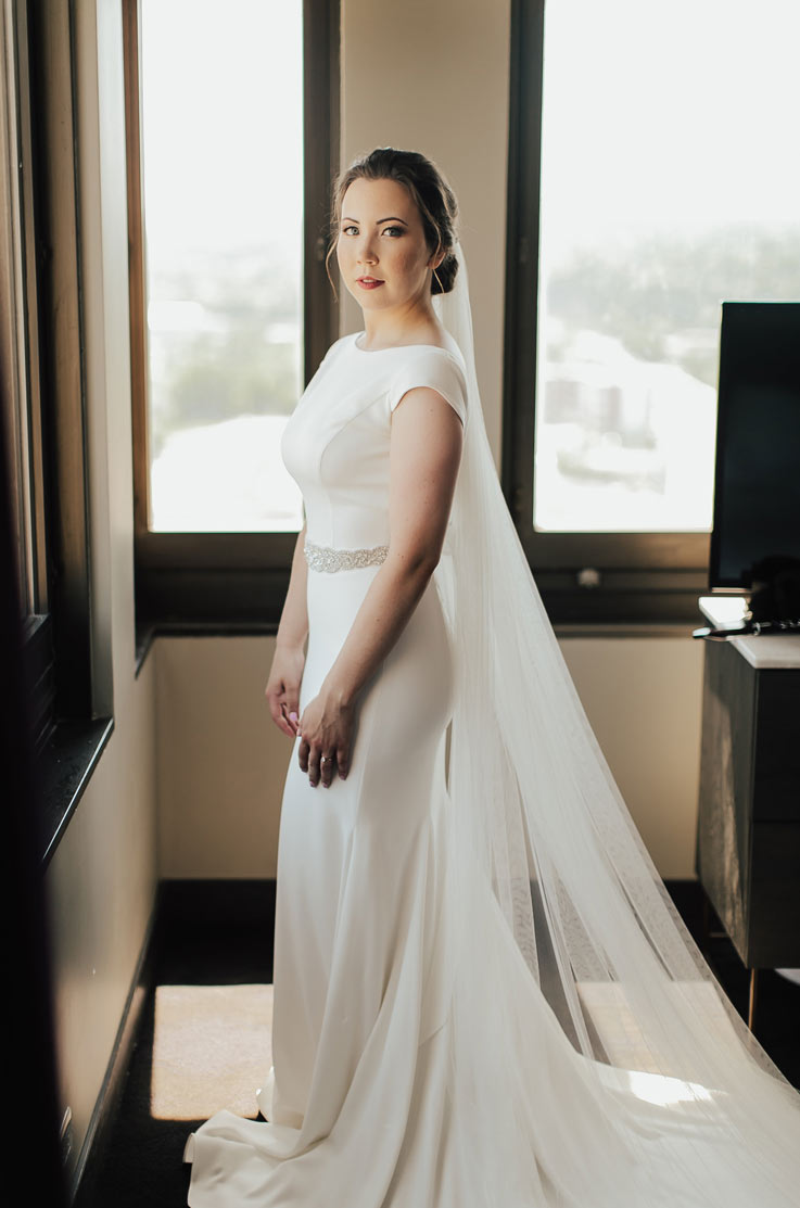 Bride stands in front of bright windows, in a crepe wedding dress with cap sleeves, a beaded belt and veil in a hotel room.