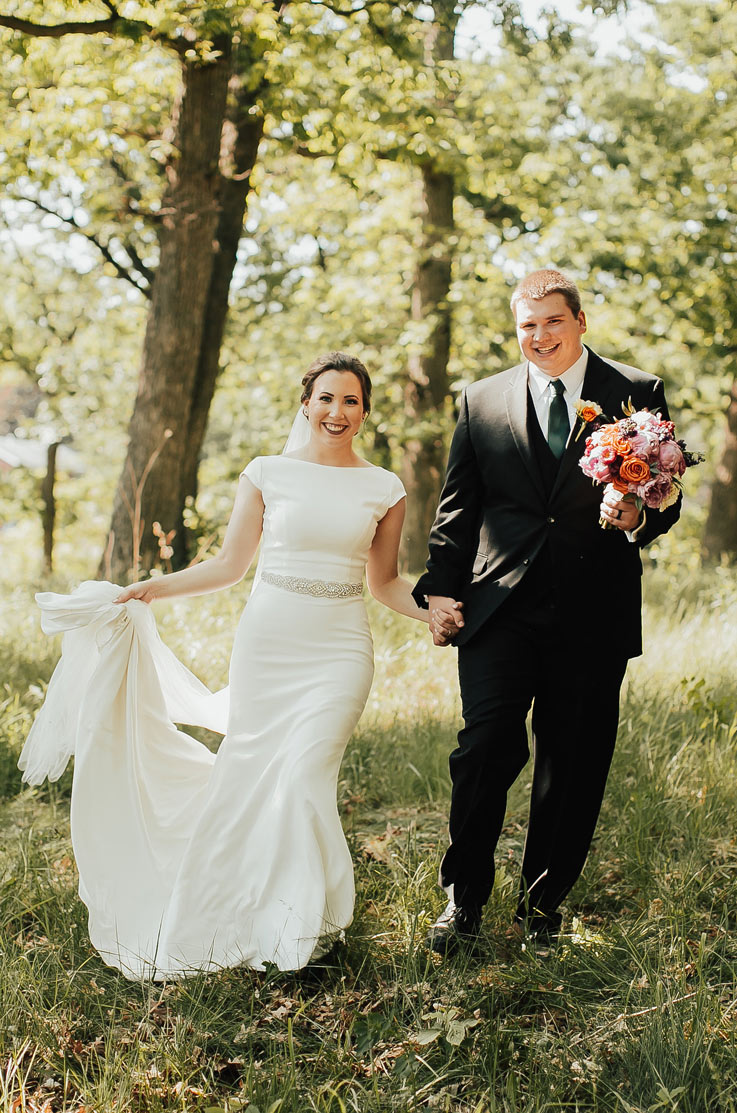  Bride in a fit and flare crepe gown with cap sleeves and belt detail, smiling holding hands with the groom in a sunny meadow. 