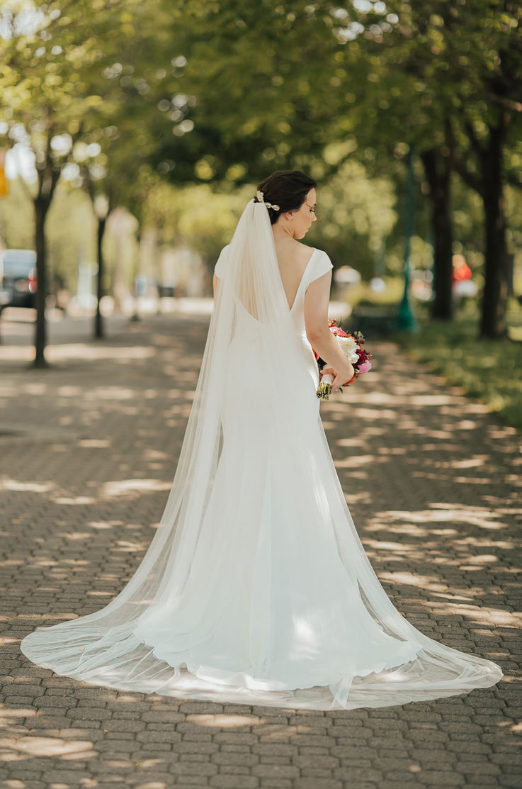 Back of bride standing on a tree-lined path, in a low back crepe gown with cap sleeves, a chapel train and a flowing veil. 